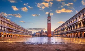 La piazza San Marco s'éveille sous un ciel aux teintes orangées. La basilique et le campanile se dressent majestueusement, reflétés sur le sol mouillé.
