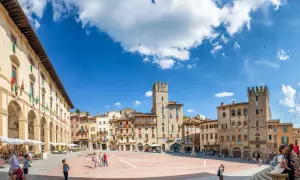 La Piazza Grande d'Arezzo, avec ses tours médiévales et ses arcades, sous un ciel bleu. Ici, la vie est belle, rythmée par les pas des passants.