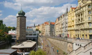 Vue de Prague que voir : tour de pierre avec dôme vert, maisons colorées le long de la rivière, et une péniche.