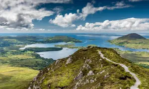 Vue panoramique du Connemara depuis Galway. Un sentier serpente sur une colline verdoyante, menant à des lacs bleus et des îles sous un ciel nuageux.