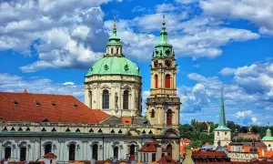 Vue imprenable sur l'église Saint-Nicolas de Prague, avec ses dômes verts et sa tour d'horloge, sous un ciel bleu et nuageux.
