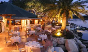 Terrasse illuminée d'un restaurant pieds dans l'eau, palmiers et tables dressées. L'endroit idéal pour un dîner au meilleur restaurant Porto Vecchio.