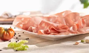 Mortadelle fine, aux éclats de pistache, évoquant son origine italienne. Une tomate et des grains de poivre complètent la scène.