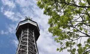 Tour Petrin, une tour d'observation métallique avec une structure en treillis, se dresse sous un ciel bleu parsemé de nuages, avec des branches d'arbre verdoyantes au premier plan.