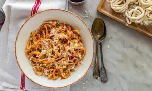 Pici all'aglione, pâtes toscanes savoureuses, servies dans un bol avec du fromage râpé. À côté, des pâtes crues enroulées.
