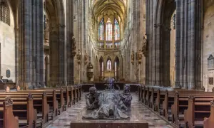 Intérieur de la cathédrale Saint-Guy au **château de Prague**. Une sculpture de bronze d'un évêque et deux moines trône au centre, face aux bancs de bois et aux vitraux colorés.