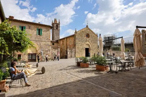 Place pavée de Monteriggioni, Italie, avec une église médiévale et des tables de café.