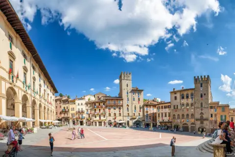 La Piazza Grande d'Arezzo, avec ses tours médiévales et ses arcades, sous un ciel bleu. Ici, la vie est belle, rythmée par les pas des passants.