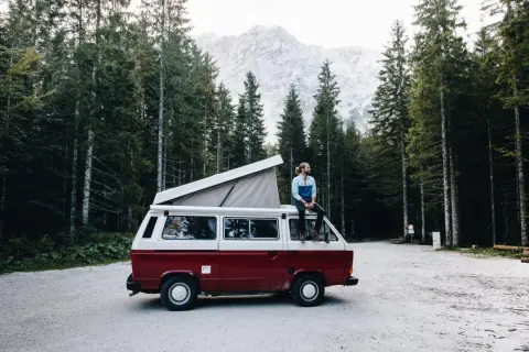 Un homme profite d'une pause lors de son tour d'Europe en van, assis sur le toit, entouré d'une forêt de pins et de montagnes majestueuses.