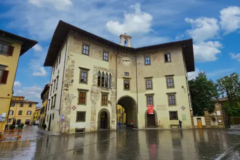 La Piazza dei Cavalieri à Pise, avec ses bâtiments historiques aux façades ocre et jaune, sous un ciel bleu et nuageux. Le sol mouillé reflète les édifices.