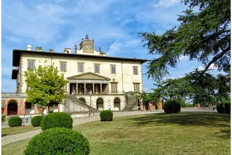 La villa Poggio a Caiano, un joyau architectural, se dresse sous un ciel bleu. Des arbres majestueux ombragent la pelouse verdoyante et les buissons taillés.