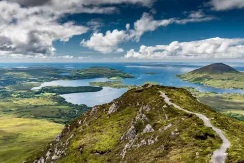 Vue panoramique du Connemara depuis Galway. Un sentier serpente sur une colline verdoyante, menant à des lacs bleus et des îles sous un ciel nuageux.