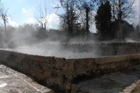 Bains antiques en pierre à San Casciano dei Bagni, dégageant une vapeur d'eau chaude sous un ciel bleu.