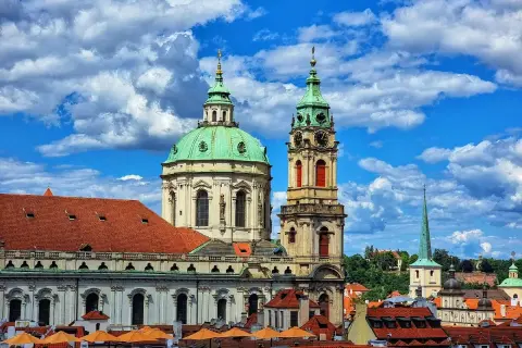 Vue imprenable sur l'église Saint-Nicolas de Prague, avec ses dômes verts et sa tour d'horloge, sous un ciel bleu et nuageux.