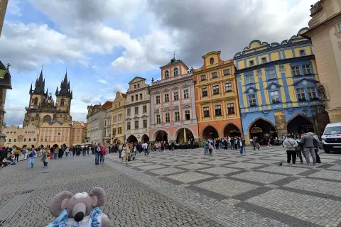 Une souris en peluche explore la place de la Vieille Ville de Prague, avec la tour de l'église Notre-Dame devant le Týn en arrière-plan.