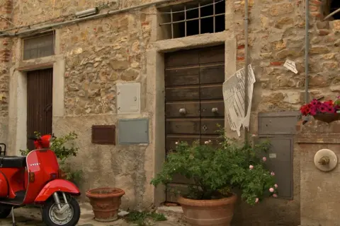 Un scooter rouge attend devant une porte en bois à Castagneto Carducci. Des fleurs roses et rouges décorent la scène.