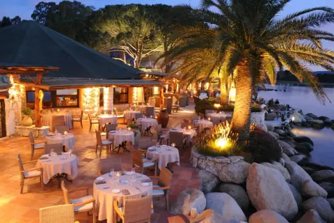 Terrasse illuminée d'un restaurant pieds dans l'eau, palmiers et tables dressées. L'endroit idéal pour un dîner au meilleur restaurant Porto Vecchio.