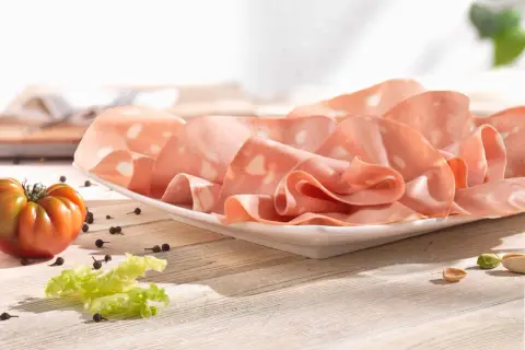Mortadelle fine, aux éclats de pistache, évoquant son origine italienne. Une tomate et des grains de poivre complètent la scène.