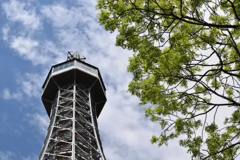Tour Petrin, une tour d'observation métallique avec une structure en treillis, se dresse sous un ciel bleu parsemé de nuages, avec des branches d'arbre verdoyantes au premier plan.
