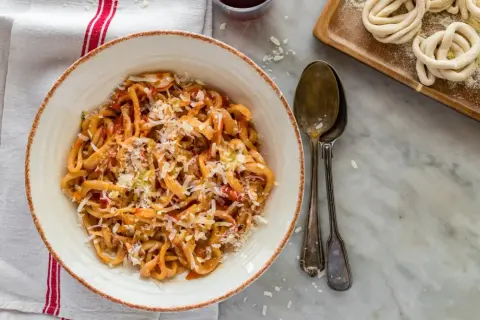 Pici all'aglione, pâtes toscanes savoureuses, servies dans un bol avec du fromage râpé. À côté, des pâtes crues enroulées.