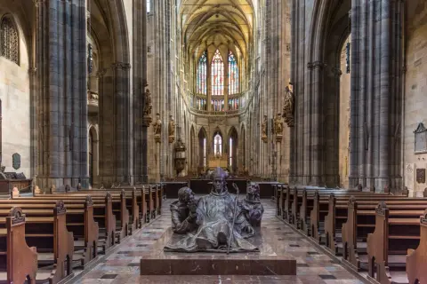 Intérieur de la cathédrale Saint-Guy au **château de Prague**. Une sculpture de bronze d'un évêque et deux moines trône au centre, face aux bancs de bois et aux vitraux colorés.