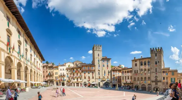 La Piazza Grande d'Arezzo, avec ses tours médiévales et ses arcades, sous un ciel bleu. Ici, la vie est belle, rythmée par les pas des passants.