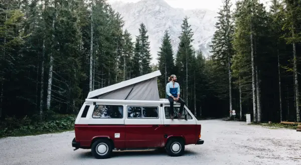 Un homme profite d'une pause lors de son tour d'Europe en van, assis sur le toit, entouré d'une forêt de pins et de montagnes majestueuses.