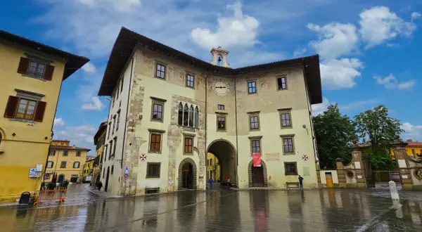 La Piazza dei Cavalieri à Pise, avec ses bâtiments historiques aux façades ocre et jaune, sous un ciel bleu et nuageux. Le sol mouillé reflète les édifices.