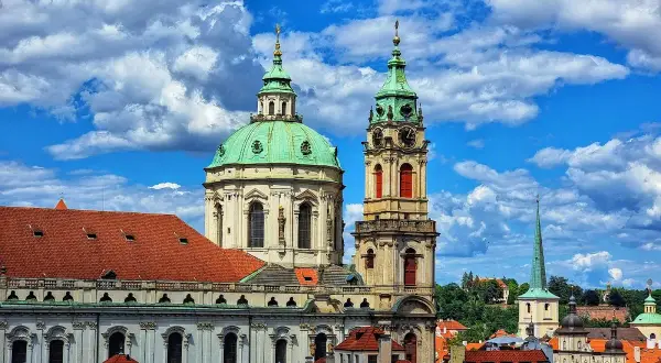 Vue imprenable sur l'église Saint-Nicolas de Prague, avec ses dômes verts et sa tour d'horloge, sous un ciel bleu et nuageux.