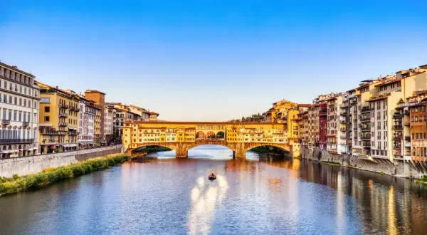 Le Ponte Vecchio, avec ses boutiques colorées, enjambe l'Arno sous un ciel bleu. Une petite barque glisse sur l'eau.