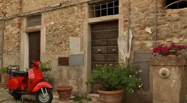 Un scooter rouge attend devant une porte en bois à Castagneto Carducci. Des fleurs roses et rouges décorent la scène.