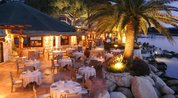 Terrasse illuminée d'un restaurant pieds dans l'eau, palmiers et tables dressées. L'endroit idéal pour un dîner au meilleur restaurant Porto Vecchio.