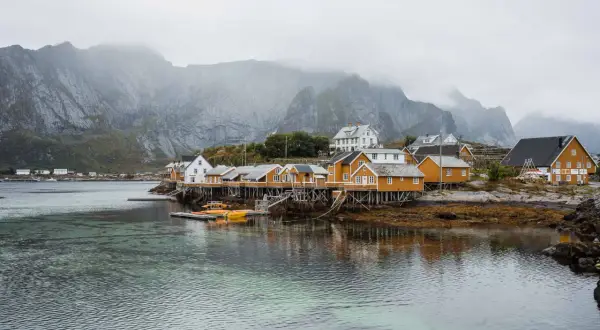 Maisonnettes jaunes sur pilotis, reflets dans l'eau, montagnes brumeuses en arrière-plan. Idéal pour un budget voyage îles Lofoten.