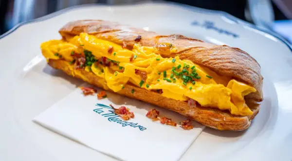 Sandwich aux œufs brouillés et bacon, parfait pour savoir où manger à Florence. Servi sur une assiette blanche avec une serviette "La Ménagère".