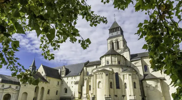 Le majestueux **château de Fontevraud**, avec sa tour imposante et ses arcs romans, se dresse sous un ciel bleu parsemé de nuages, encadré par le feuillage verdoyant des arbres.