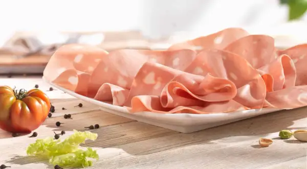 Mortadelle fine, aux éclats de pistache, évoquant son origine italienne. Une tomate et des grains de poivre complètent la scène.
