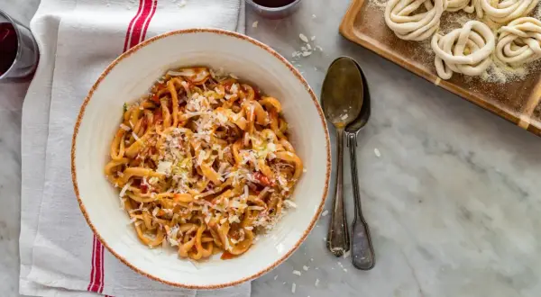 Pici all'aglione, pâtes toscanes savoureuses, servies dans un bol avec du fromage râpé. À côté, des pâtes crues enroulées.