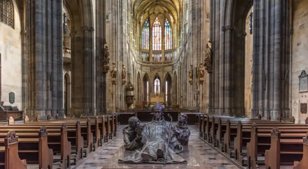 Intérieur de la cathédrale Saint-Guy au **château de Prague**. Une sculpture de bronze d'un évêque et deux moines trône au centre, face aux bancs de bois et aux vitraux colorés.