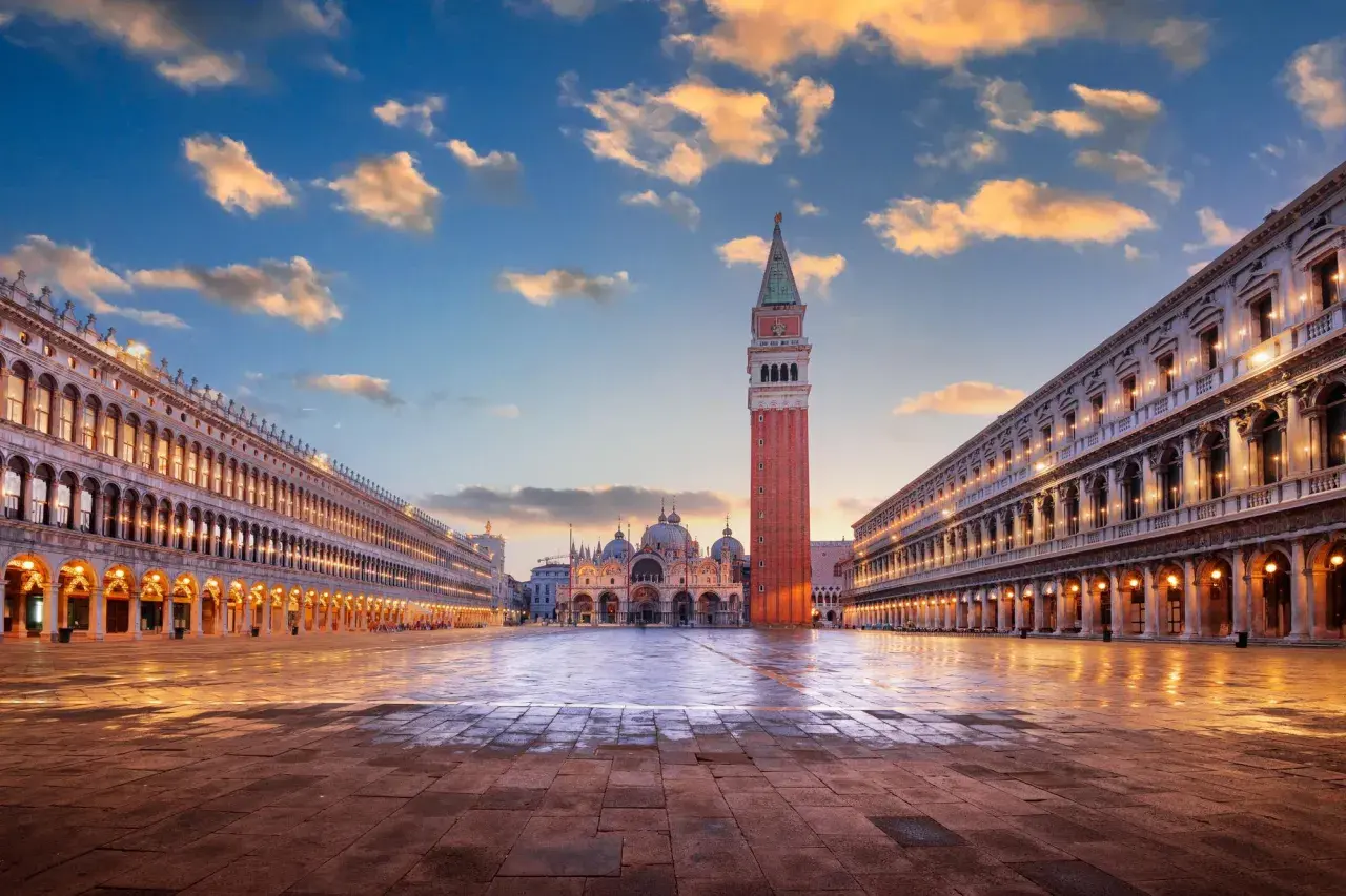 La piazza San Marco s'éveille sous un ciel aux teintes orangées. La basilique et le campanile se dressent majestueusement, reflétés sur le sol mouillé.