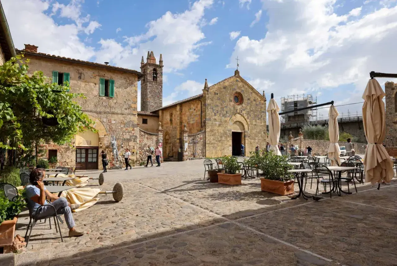 Place pavée de Monteriggioni, Italie, avec une église médiévale et des tables de café.