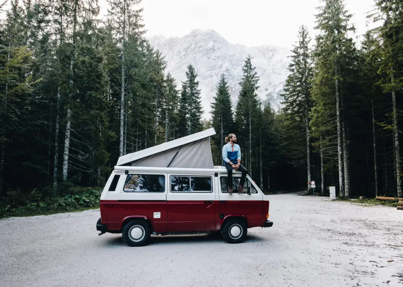 Un homme profite d'une pause lors de son tour d'Europe en van, assis sur le toit, entouré d'une forêt de pins et de montagnes majestueuses.