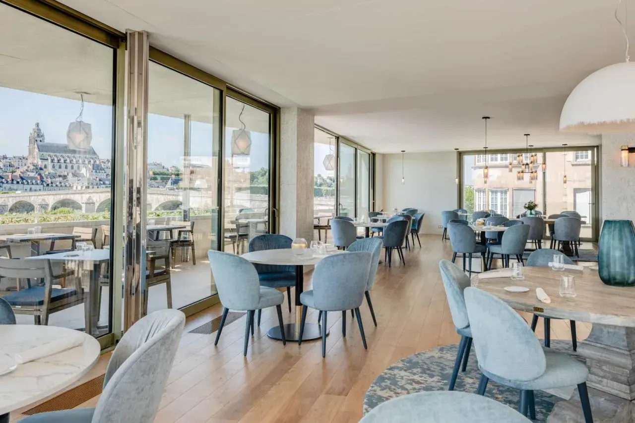 Salle lumineuse avec vue panoramique sur la Loire. Tables dressées, chaises bleues confortables. Un cadre idéal pour découvrir les restaurants de la Loire.