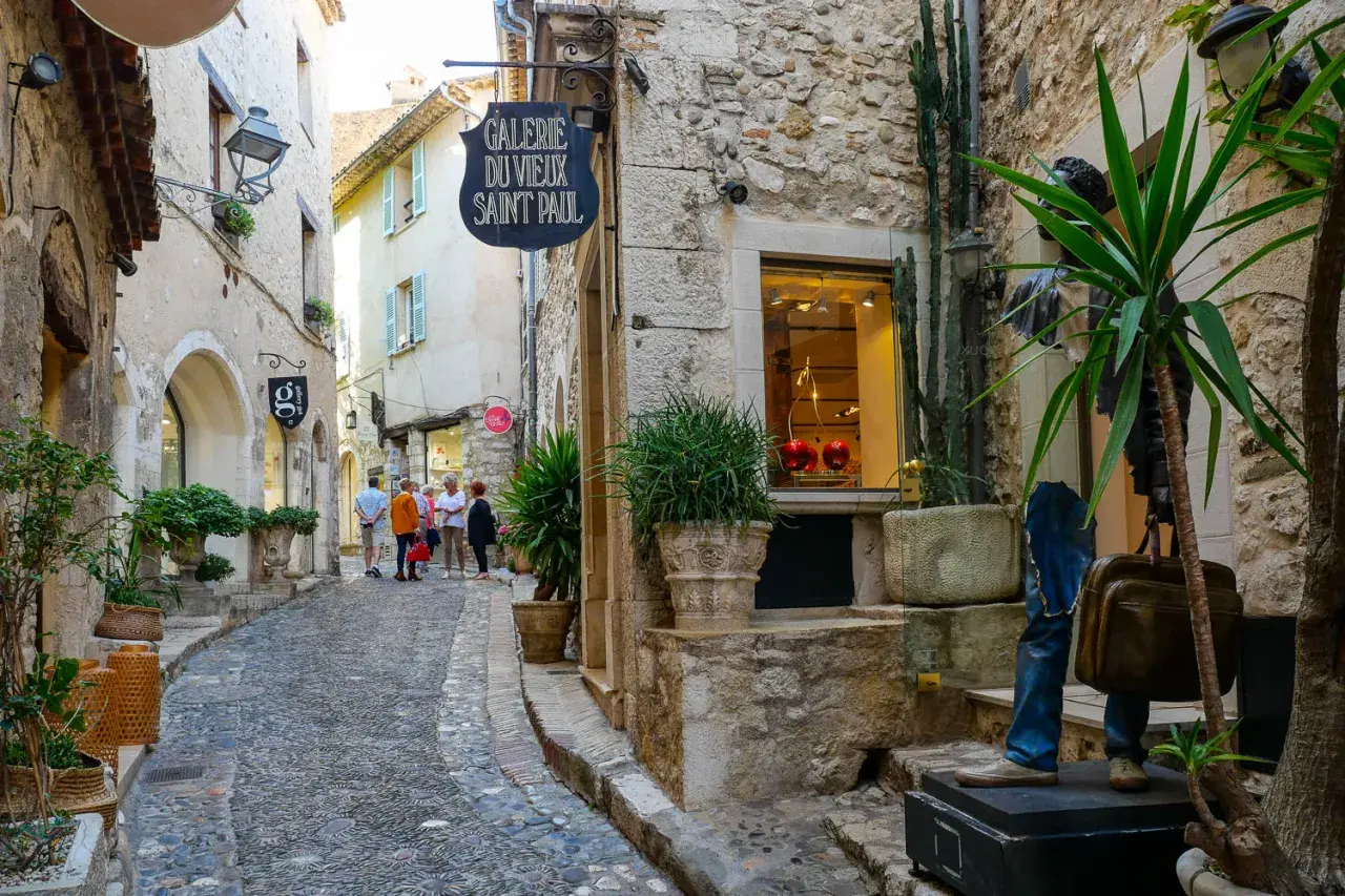 Ruelle pavée à Saint-Paul-de-Vence, avec des boutiques, des plantes et des touristes.