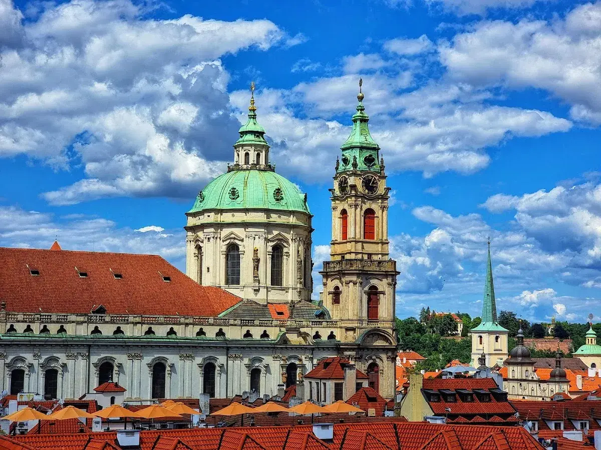 Vue imprenable sur l'église Saint-Nicolas de Prague, avec ses dômes verts et sa tour d'horloge, sous un ciel bleu et nuageux.