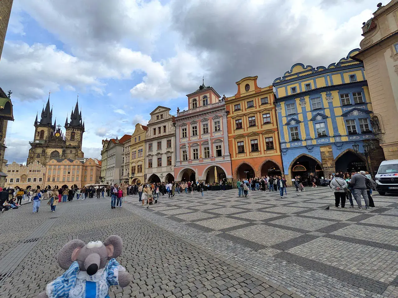 Une souris en peluche explore la place de la Vieille Ville de Prague, avec la tour de l'église Notre-Dame devant le Týn en arrière-plan.