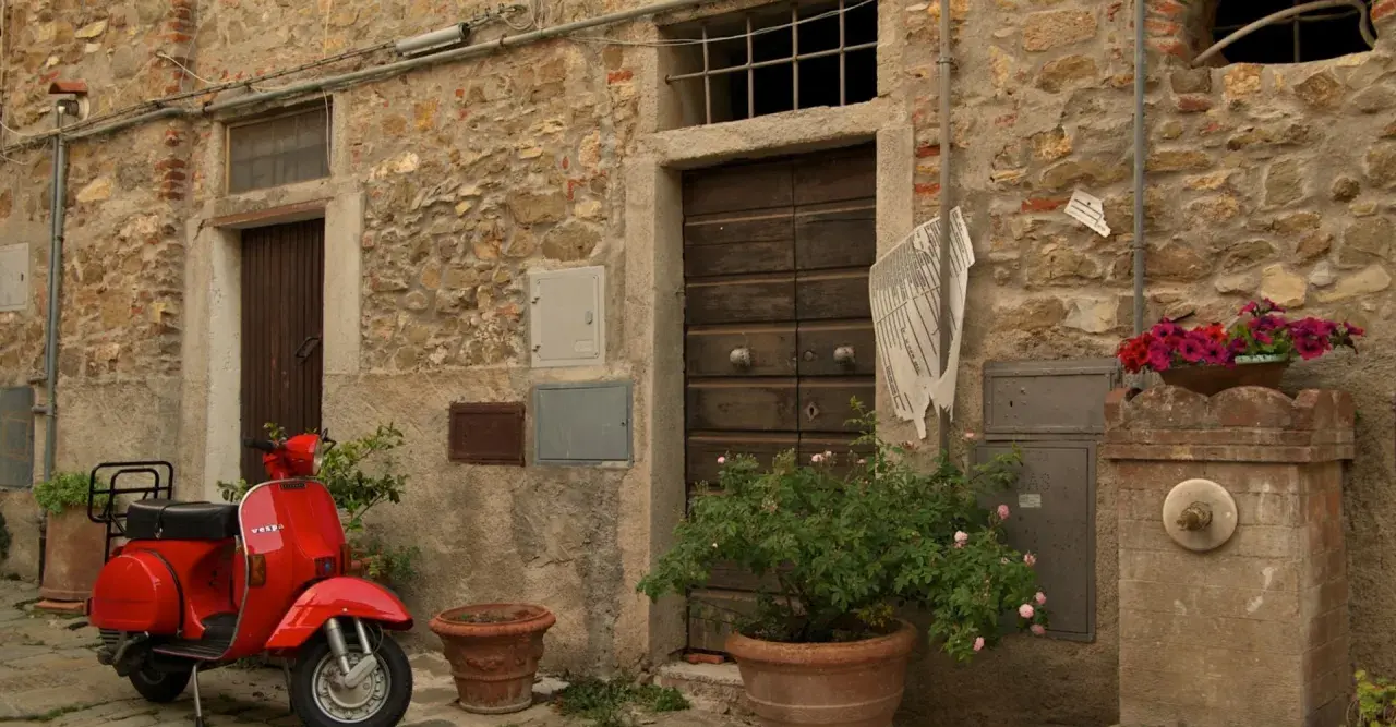 Un scooter rouge attend devant une porte en bois à Castagneto Carducci. Des fleurs roses et rouges décorent la scène.