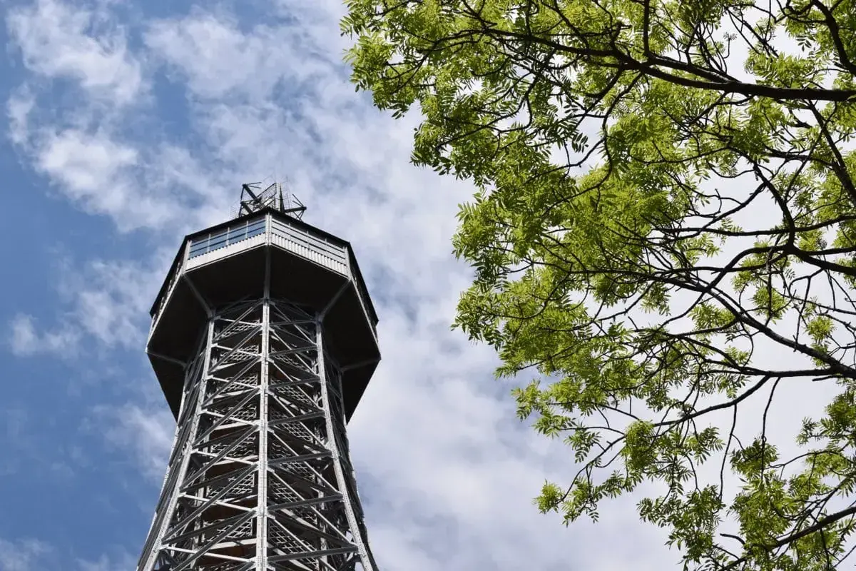 Tour Petrin, une tour d'observation métallique avec une structure en treillis, se dresse sous un ciel bleu parsemé de nuages, avec des branches d'arbre verdoyantes au premier plan.