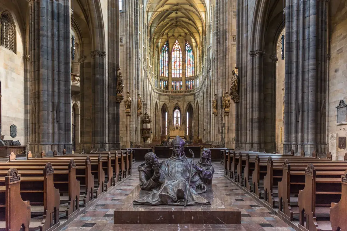 Intérieur de la cathédrale Saint-Guy au **château de Prague**. Une sculpture de bronze d'un évêque et deux moines trône au centre, face aux bancs de bois et aux vitraux colorés.