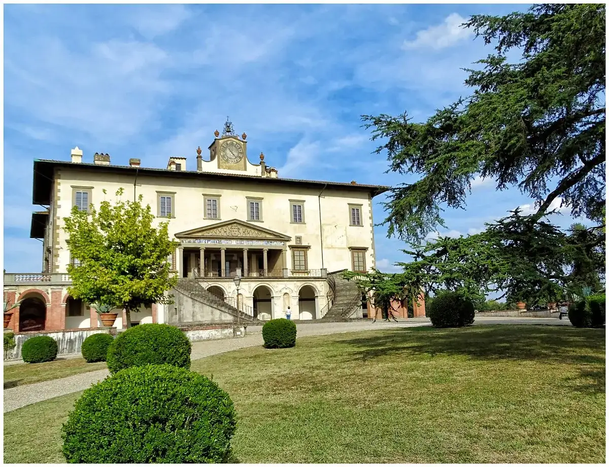 La villa Poggio a Caiano, un joyau architectural, se dresse sous un ciel bleu. Des arbres majestueux ombragent la pelouse verdoyante et les buissons taillés.