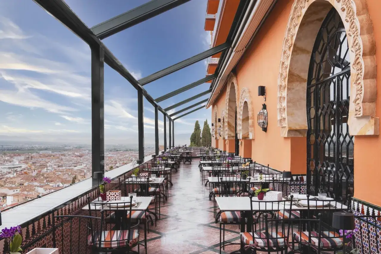 Terrasse d'un restaurant à Grenade, avec vue panoramique sur la ville. Tables dressées sous une structure métallique et un ciel bleu.